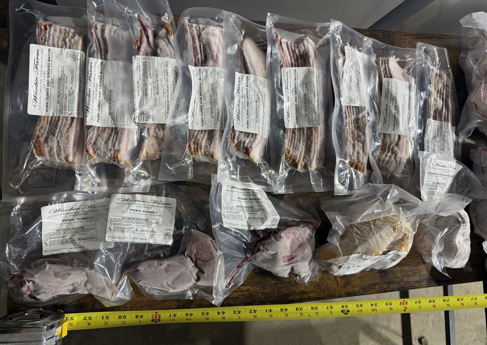 Vacuum-sealed packages of pork cured and smoked bacon labeled Woobie Farm, lined up next to a tape measure